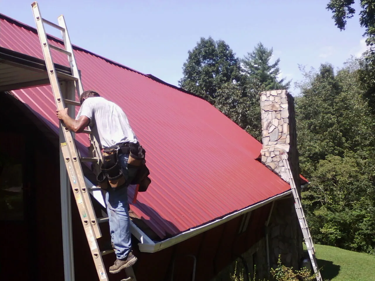 Roofer on a ladder beside a red metal roof and stone chimney — Soffit & Fascia Repair by Roofing Brothers in Salineville